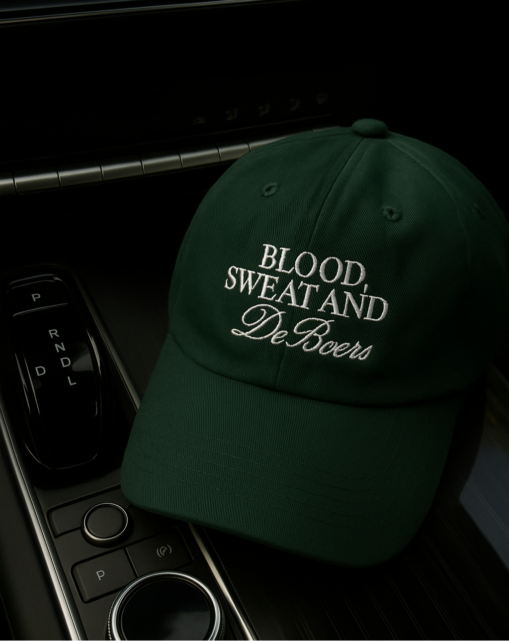 Close up of ivy green women’s baseball cap on car dash, featuring embroidered text: “Blood, Sweat & DeBeers”