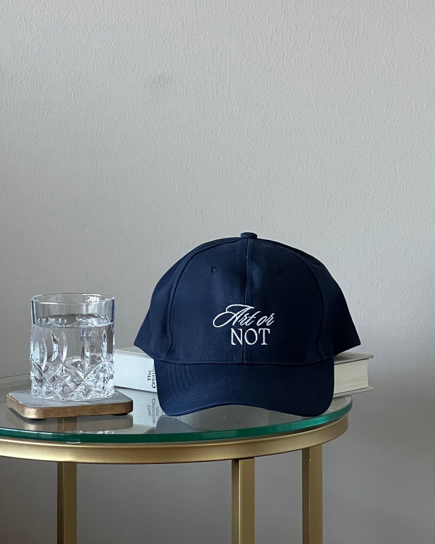 Close up of navy blue baseball cap on luxurious bedside table with crystal water glass, featuring embroidered phrase: “Art or Not”