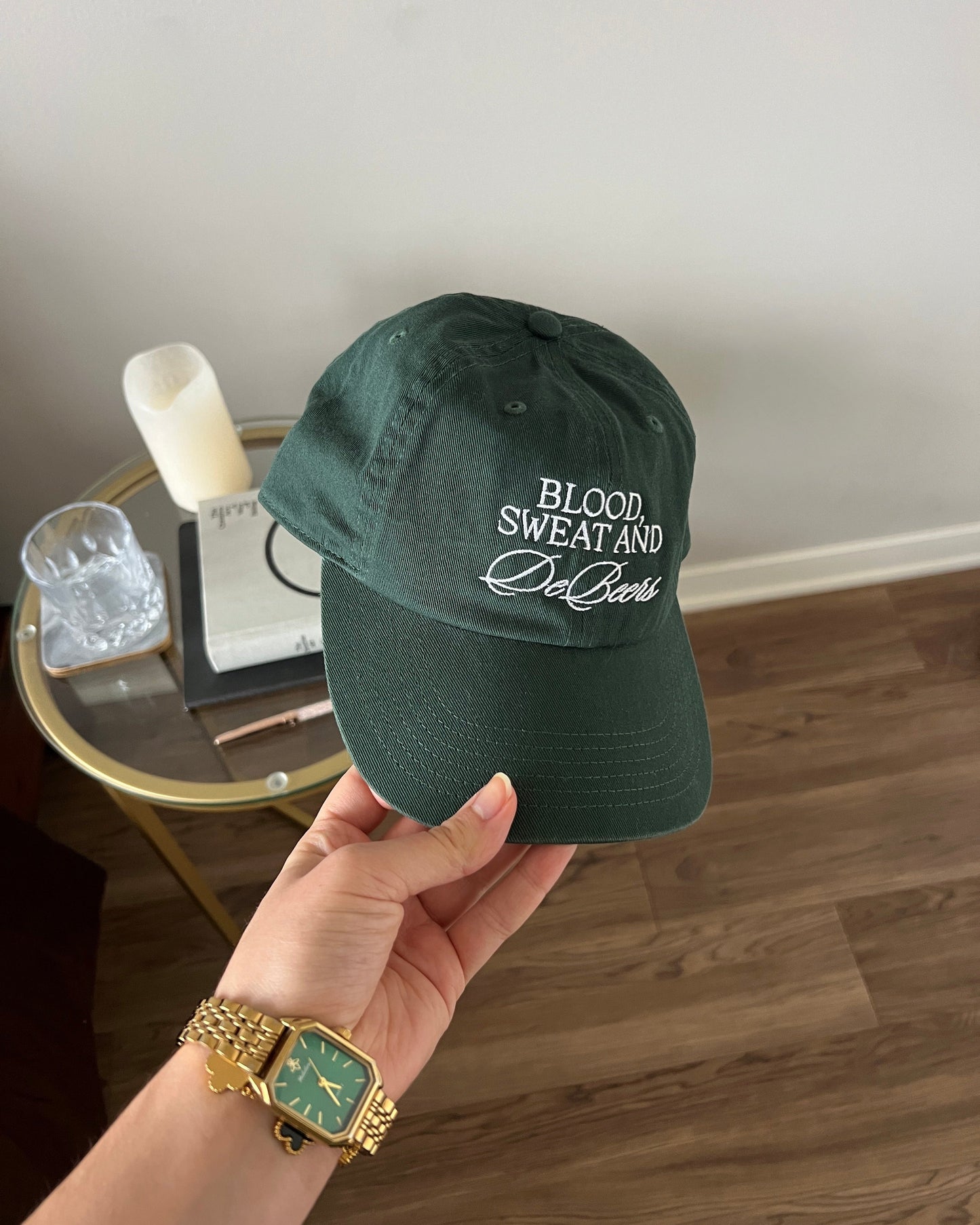 Woman holding ivy green women’s baseball cap, featuring luxury-inspired embroidered text: “Blood, Sweat & DeBeers”