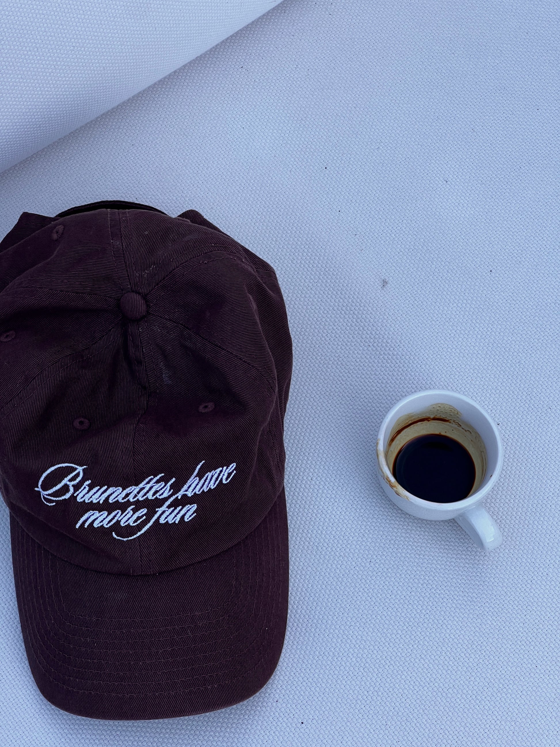 Brown embroidered baseball cap with “Brunettes Have More Fun” next to an espresso cup