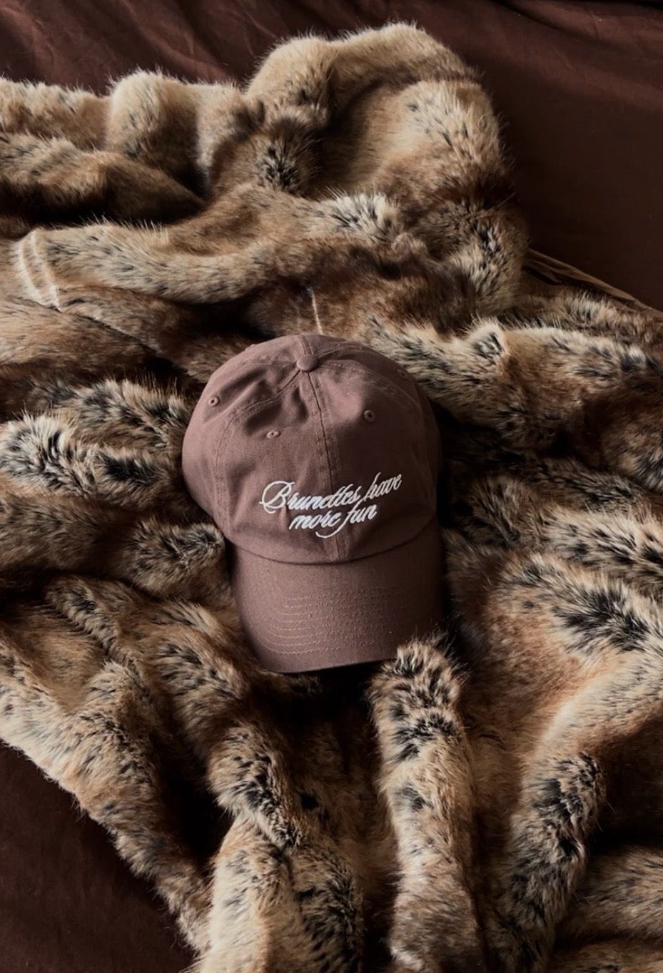 Brown embroidered baseball cap with “Brunettes Have More Fun” styled on a fur throw